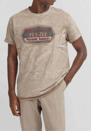 Uomo che indossa una camicia beige a maniche corte e pantaloni, camicia con scritta "YES ZEE Premium Garment" e grafica sul petto, mano in tasca.