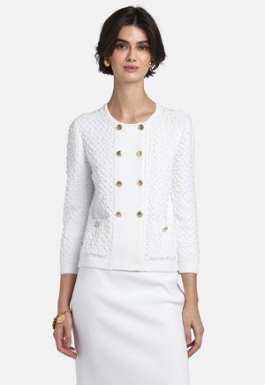 Vrouw met een witte textuurachtige jas met gouden knoppen en voorzakken over een witte jurk, geaccentueerd met gouden oorbellen en een armband.