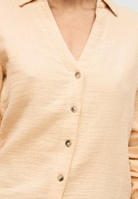 Chemise en coton beige texturé avec un large col et quatre boutons marron visibles sur le devant, portée par une personne.