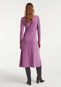 Vestido largo de manga larga en color púrpura con un corsé ajustado y falda acampanada, hecho de una tela suave. Combinado con botas negras hasta la rodilla.