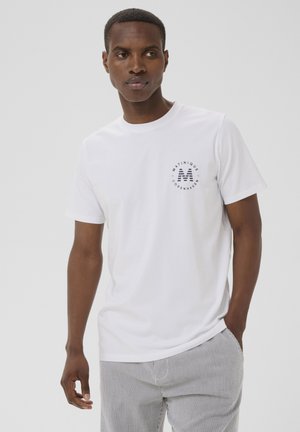 Homme portant un T-shirt blanc avec le logo "Matinique Copenhagen" et un pantalon rayé, debout avec une main dans la poche devant un fond uni.