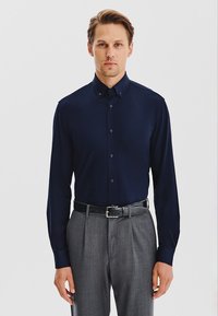 Granatowa koszula z długim rękawem z kołnierzykiem zapinanym na guziki, miękka w dotyku, z guzikami z przodu. Połączona z szarymi spodniami i czarnym paskiem.