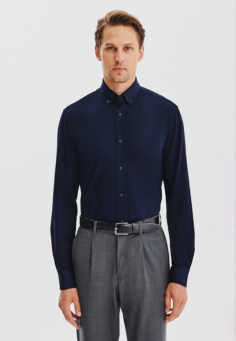 Granatowa koszula z długim rękawem z kołnierzykiem zapinanym na guziki, miękka w dotyku, z guzikami z przodu. Połączona z szarymi spodniami i czarnym paskiem.