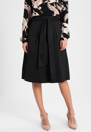 Femme portant une jupe noire au genou avec une ceinture nouée à la taille, associée à des chaussures à talons hauts couleur chair et un chemisier noir avec un imprimé grue blanche.