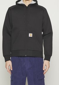 Zwarte zip-up hoodie met een grijze capuchonvoering, voorkruiken, ribbels bij de mouwen en een Carhartt logo patch op de linkerzak.