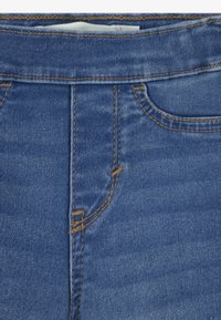 Primer plano de la cintura de unos jeans de mezclilla azules con costuras naranjas y una etiqueta blanca de ropa visible metida dentro.