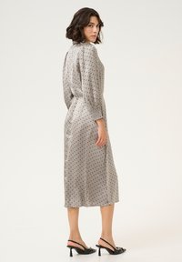 Grijze jurk met stippen, een middellange ronde halslijn, opgezette mouwen en een getailleerde taille. Draagt zwarte slingback hakken.