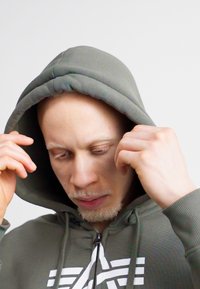 Zelená mikina s kapucňou a šnúrkou, s klokaním vreckom, obsahujúca biele grafické detaily na hrudníku. Tkanina vyzerá jemne a štruktúrovane.