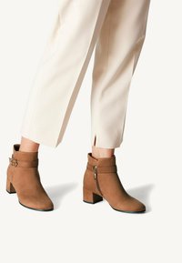 Brown suede ankle boots with a block heel, featuring a side zipper and buckle strap detail, worn with cream trousers.