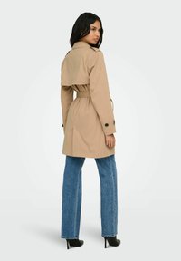 Femme portant un trench-coat beige ceinturé, un jean bleu et des bottes à talons noirs, debout et regardant par-dessus son épaule.