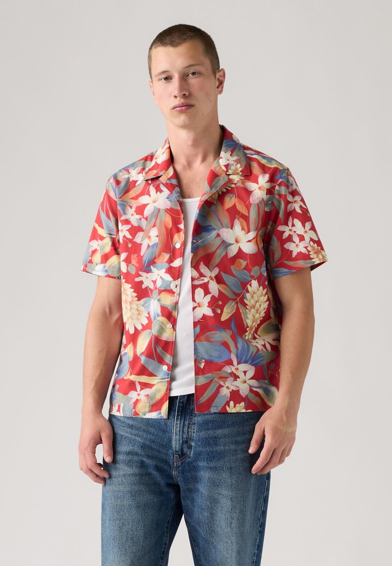 Joven con camisa roja de manga corta con estampado floral blanco, camiseta blanca y vaqueros azules sobre un fondo liso.