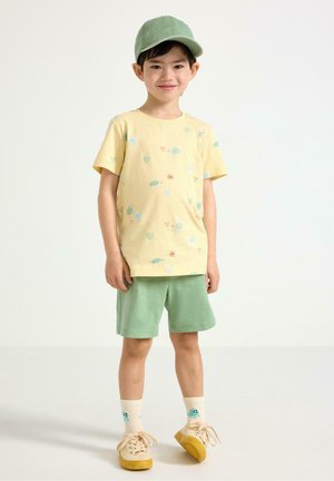 Ung dreng stående iført grøn kasket, gul blomstret t-shirt, grønne shorts, beige sko og cremefarvede sokker med blå ansigtsdesigns.