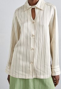 Camicia button-up color crema con righe verticali marroni, colletto appuntito e dettaglio a chiave sullo scollo. Tessuto in cotone morbido.