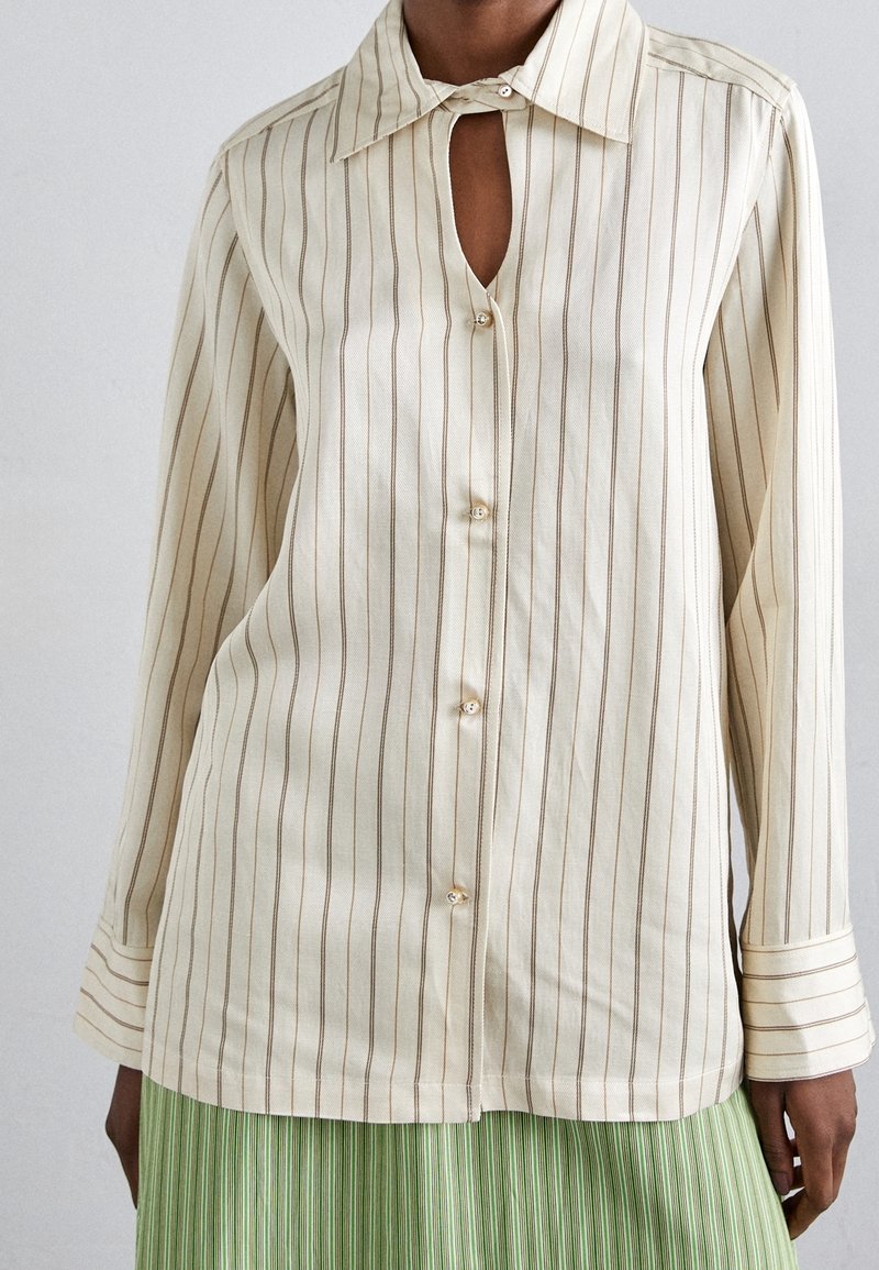 Camicia button-up color crema con righe verticali marroni, colletto appuntito e dettaglio a chiave sullo scollo. Tessuto in cotone morbido.
