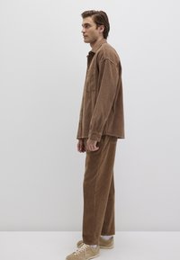 Braunes Kordhemd mit Knöpfen und Brusttasche, kombiniert mit passenden braunen Kordhosen. Getragen mit hellen Sneakers.