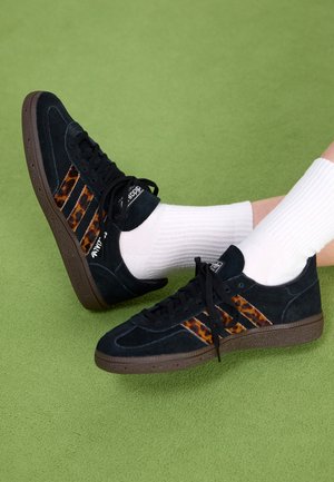 Baskets Adidas noires avec des rayures imprimé léopard marron portées avec des chaussettes blanches côtelées sur une surface verte.
