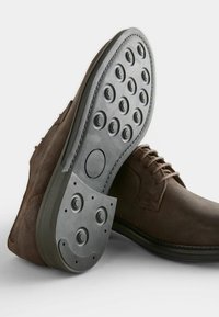 Brown suede shoes with a rubber sole featuring circular grip patterns. Lace-up design with a smooth texture and contrasting stitching.