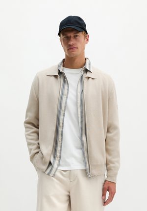 Homme portant une casquette noire, une veste beige zippée par-dessus une chemise rayée à boutons et un t-shirt blanc, les mains dans les poches devant un fond blanc.