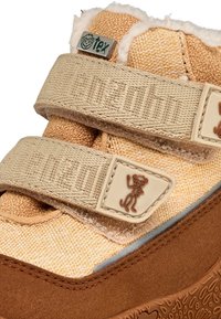 Chaussure en textile beige avec des bandes velcro, doublure douce et semelle en caoutchouc brun avec un motif antidérapant. Inclut un accent de logo ludique.