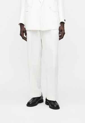 Mężczyzna w dopasowanych białych spodniach garniturowych, z dopasowaną dwurzędową marynarką i czarnymi skórzanymi mokasynami na białym tle.