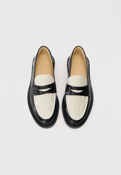 Sort og hvid læder loafers med præget design på den hvide forfod og sort rem, vist fra oven på en ensfarvet baggrund.
