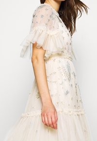 Ärmlös klänning i elfenben med genomskinliga volangärmar, intrikata silver- och blå stjärnbroderier samt en upptagen midja med volangdetaljer.