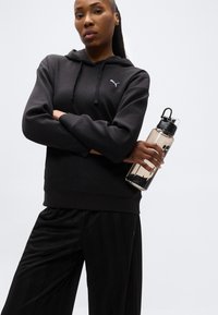 Γυναίκα με μαύρο φούτερ και παντελόνι Puma κρατάει ένα μπουκάλι νερό, στέκεται με σταυρωμένα τα χέρια μπροστά σε λευκό απλό φόντο.