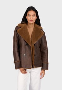 Brązowa skórzana podwójnie zapinana kurtka z brązowym kołnierzem z owczej wełny, z czarnymi guzikami i obszytymi rękawami, noszona na jasnobezowym topie.