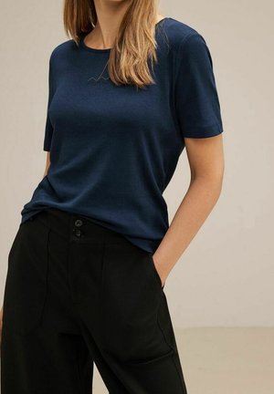 Vrouw met een marineblauw T-shirt met korte mouwen, ingestopt in een zwarte high-waist broek, met handen in de zakken, beige achtergrond.