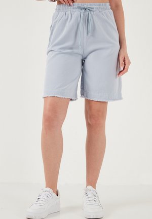 Žena stojaca proti jednoduchému pozadiu, oblečená v svetlomodrých šortkách so stiahnutím na šnúrku a orezanými lemami, a bielych teniskách.