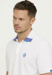 Witte polo-shirt van katoen, met een blauw logo op de borst en een kraag met blauwe en rode strepen. Korte mouwen, klassiek ontwerp.
