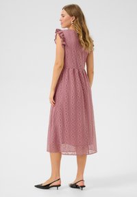 Vaaleanpunainen pitsinen midimekko, jossa on timanttikuvio, röyhelöpäät ja vyötäröliuska, yhdistettynä mustiin slingback-kengiin.