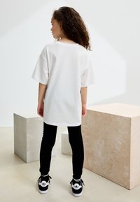 Maglietta bianca oversize a maniche corte, abbinata a leggings neri e sneakers nere con dettagli bianchi, su sfondo minimalista.