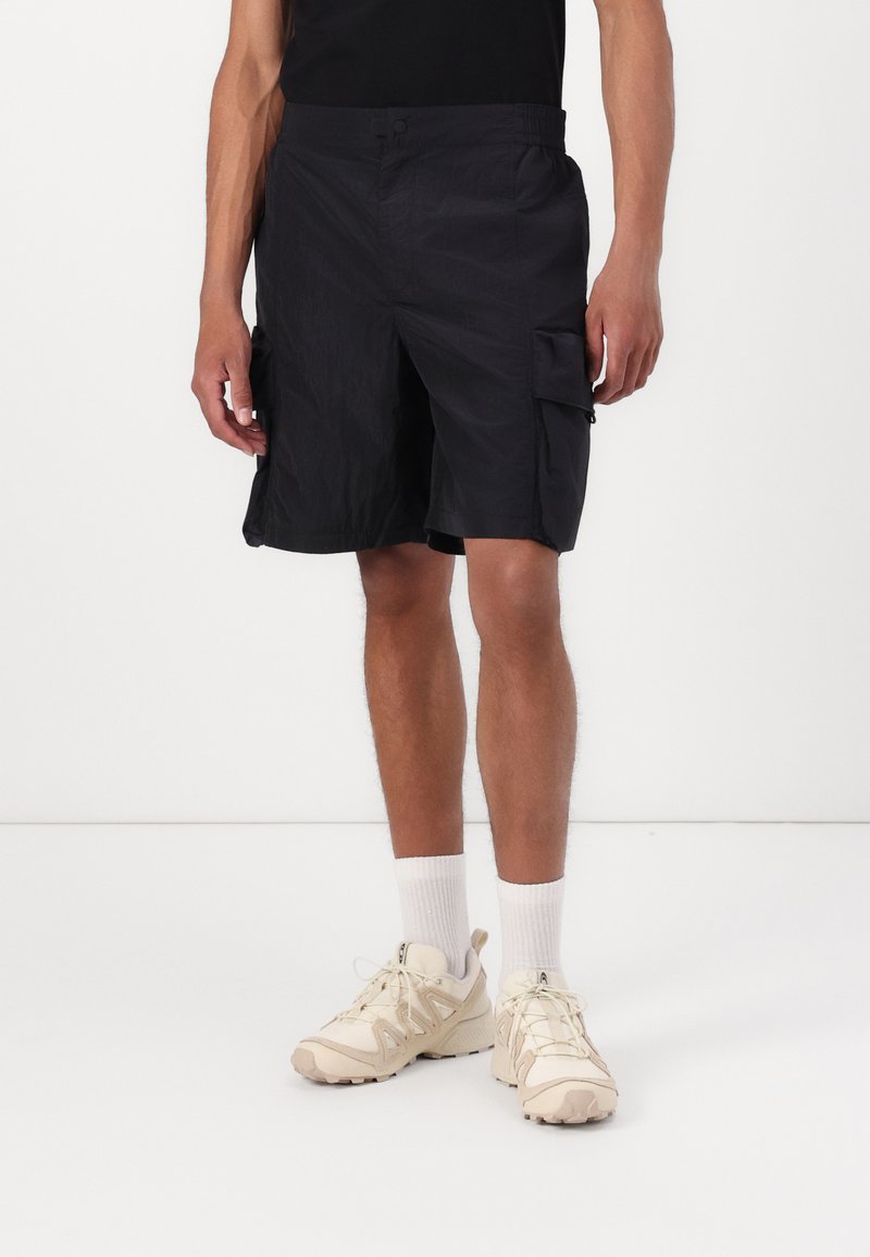 Black cargo shorts in lightweight fabric, featuring side pockets and a relaxed fit. Paired with beige sneakers and white ankle socks.