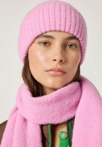 Roze gebreide muts met een geribbelde structuur, gecombineerd met een bijpassende roze sjaal met een zachte, pluizige textuur. Het model draagt een groen shirt eronder.