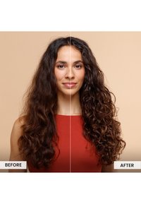 Frau mit langen braunen Haaren, die ein rotes ärmelloses Oberteil trägt, zeigt Vorher- und Nachher-Ergebnisse von Haaren, die in definierte Locken frisiert sind.