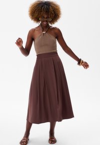 Bruine geplisseerde rok gecombineerd met een geribbelde haltertop in een lichte taupe tint. De uitstraling bevat geaccentueerde strikken en lichte sandalen.