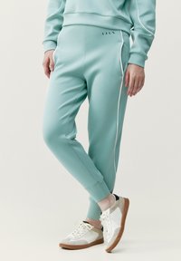 Pantaloni della tuta verde menta con piping bianco sui lati, orlo sfiancato e texture liscia. Indossati con sneakers bianche e beige dotate di lacci.