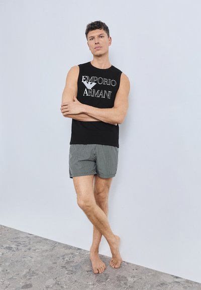 Sort ærmeløs tanktop med "Emporio Armani"-logo, kombineret med stribede svømmeshorts i grå og hvide nuancer. Model står barfodet.