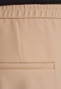 Beige stof, met textuur, met een verticale ribstructuur. Heeft een enkele horizontale pocket. Naaddetail zichtbaar aan de linkerkant.