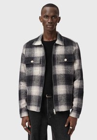Veste à carreaux en mélange de laine noir et blanc, avec un col, deux poches poitrine et une fermeture éclair à l'avant, stylée avec un pull noir et des jeans.