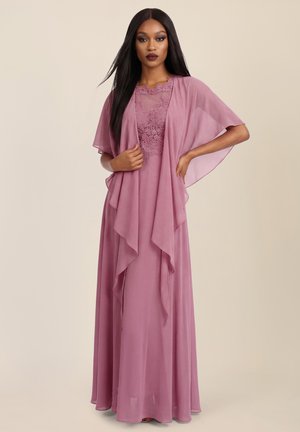 Frau trägt ein bodenlanges mauvefarbenes Kleid mit Spitzenoberteil und transparenten Umhangärmeln, steht mit einer Hand in der Hüfte und hält mit der anderen den Umhang.