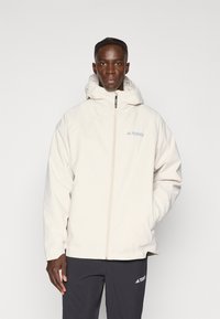 Light beige jacket with a zip closure and adjustable hood, featuring the "Terrex" logo in light blue. Smooth, waterproof fabric texture.