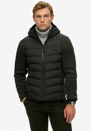 Chaqueta acolchada negra con capucha y mangas de punto gris. Cierre frontal con cremallera, logo en el pecho, detalles de tela texturizada, diseño ajustado.