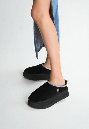 Zoccoli in suede nera con punta arrotondata e suola in gomma texturizzata. Accento argentato attorno all'apertura sulla caviglia. Design semplice e facile da indossare.