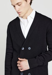 Schwarze Strickjacke mit strukturiertem Strick, doppelt geknöpft und vier blauen Knöpfen. Getragen über einem weißen Hemd mit Kragen.