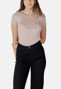 Camiseta ajustada de color rosa claro con texto metálico dorado "EA7 EMPORIO ARMANI", con cuello redondo y mangas cortas, combinada con pantalones negros.