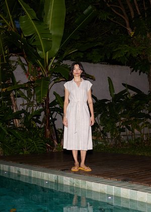 Femeie într-o rochie ușoară cu dungi și sandale galbene stând pe o terasă de lemn lângă o piscină placată cu gresie, înconjurată de plante tropicale mari, verzi, noaptea.