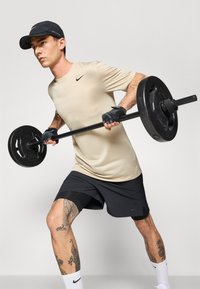 Homme portant un t-shirt beige Nike et un short noir soulevant une barre avec des poids, montrant des tatouages sur les jambes, sur fond uni.