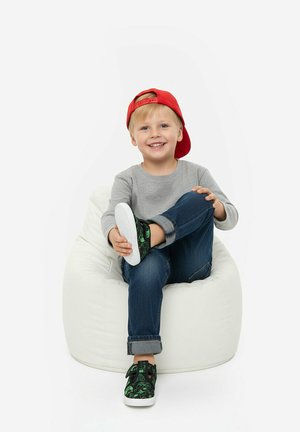 Smiling young boy wearing a red backward cap, gray shirt, blue jeans, and green sneakers sits cross-legged on a white bean bag chair.